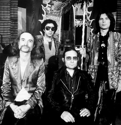 Schwarzweißfoto einer vierköpfigen Rockband mit markanter Bühnenkleidung